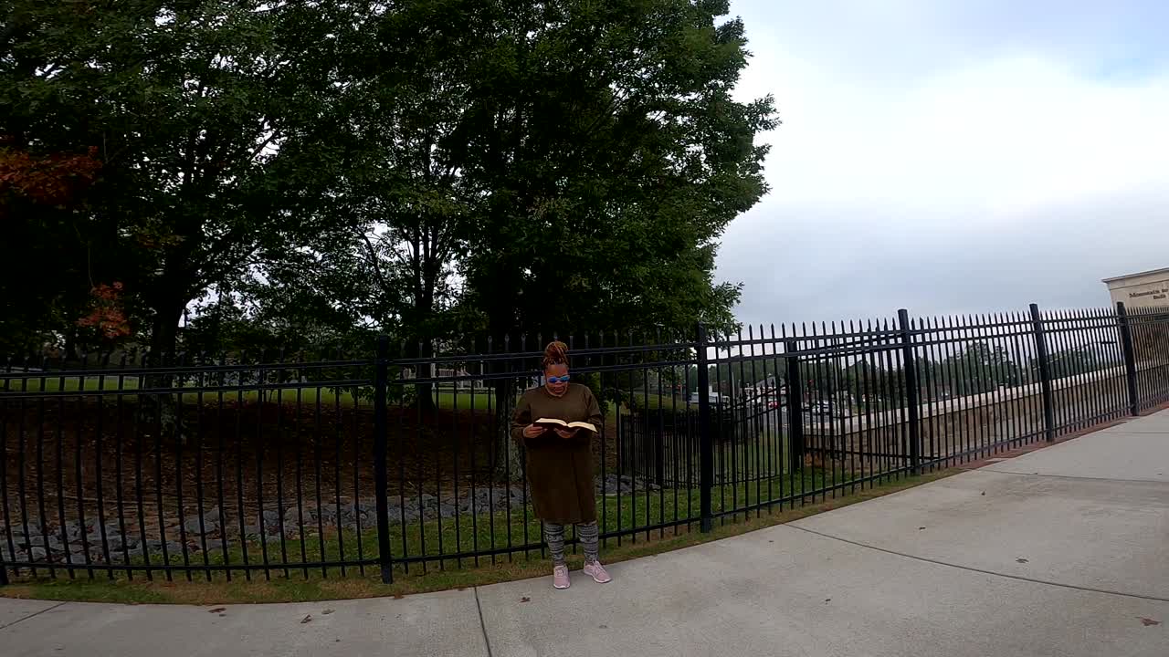 mujer negra de pie junto a la cerca de hierro en un día nublado leyendo la biblia