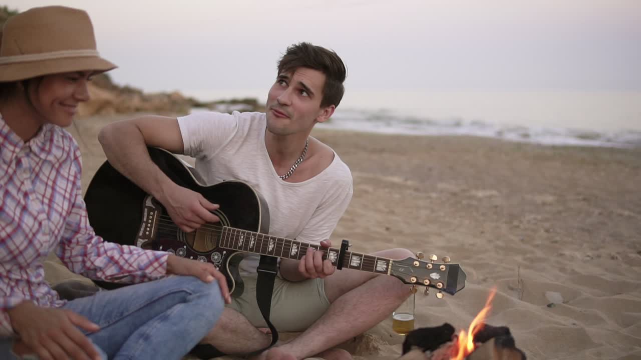 un joven está tocando la guitarra junto al fuego sentado en la playa junto con amigos. su novia trae una salchicha a la parrilla
