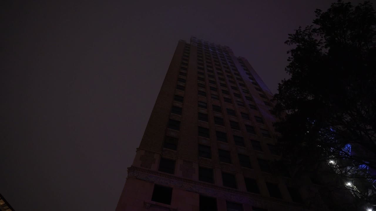 A dark, moody shot tilts upward, revealing a towering building shrouded in thick fog, its faint lights barely piercing the night