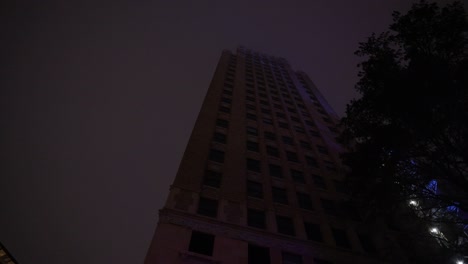 a dark, moody shot tilts upward, revealing a towering building shrouded in thick fog, its faint lights barely piercing the night