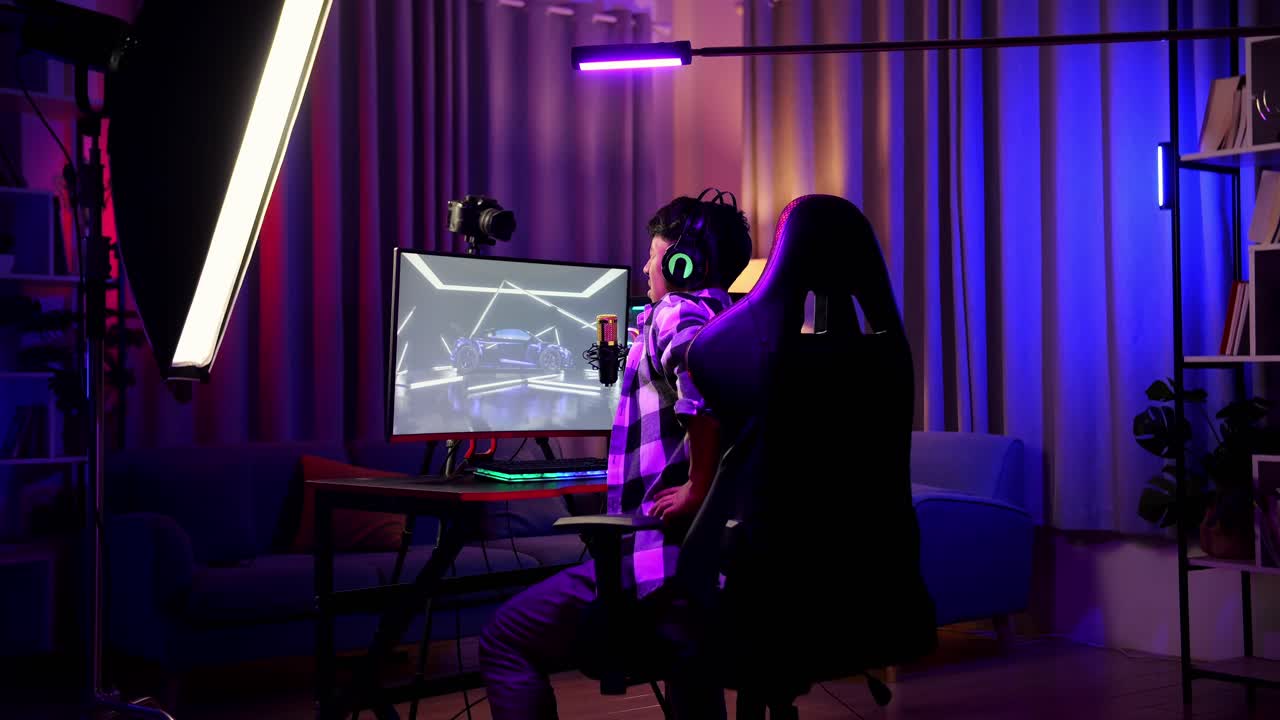 개인 컴퓨터에서 네트워크를 통해 자동차 경주 게임을하는 동안 허리 통증이있는 아시아 소년 스트리머의 뒷면. 라이브 스트리밍 비디오 게임, rgb led 스트립 라이트에 의해 조명 된 책상