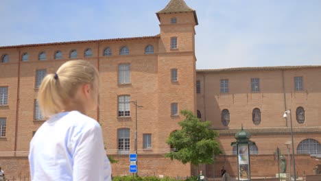 a blonde caucasian woman walks down the street in front of a big old red stone building with lots of windows