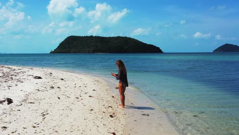 Attractive-young-woman-walking-on-beautiful-exotic-beach-washed-by-calm-clear-transparent-sea-water-and-taking-selfie-photos-in-Thailand