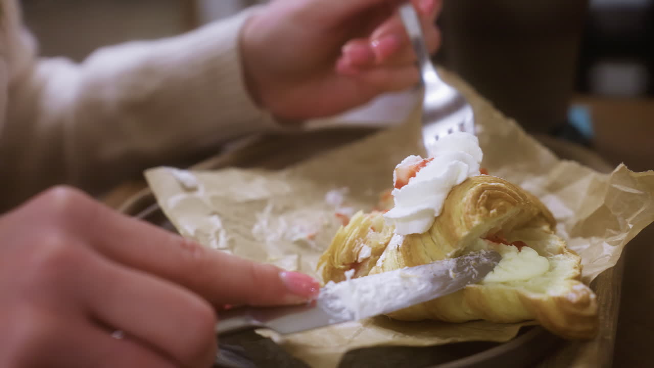 Close-up shot of female hand using fork to dig into creamy croissant topped with whipped cream and fruit. Croissant cut into with knife in cozy cafe, blurred background
