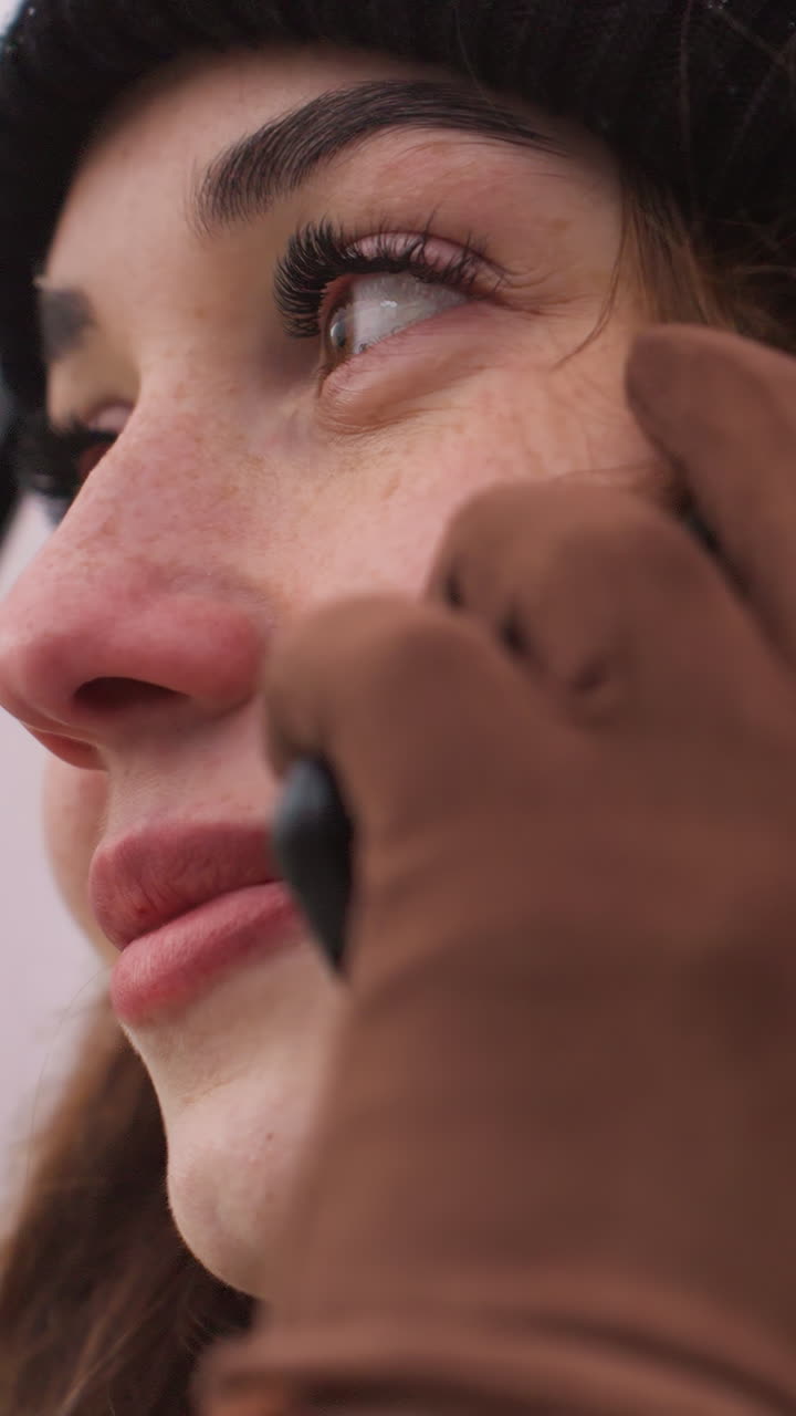 Close-up side view of lady in black cap, brown jacket, and gloves, holding black phone to her ear, with a blurred background of nature and urban elements