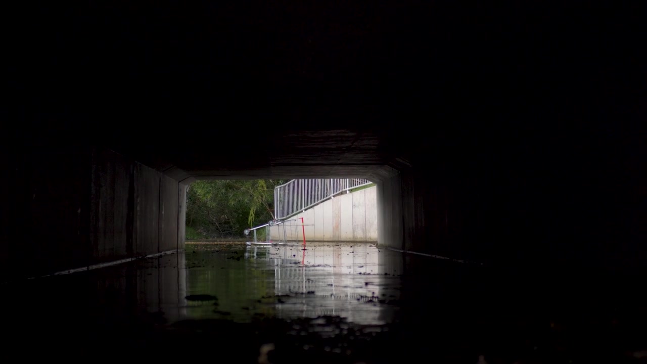 Slow motion landscape of a discarded shopping trolley tipped over inside a concrete road underpass and dark tunnel drainage system with water residue on the ground and metal railings in the background