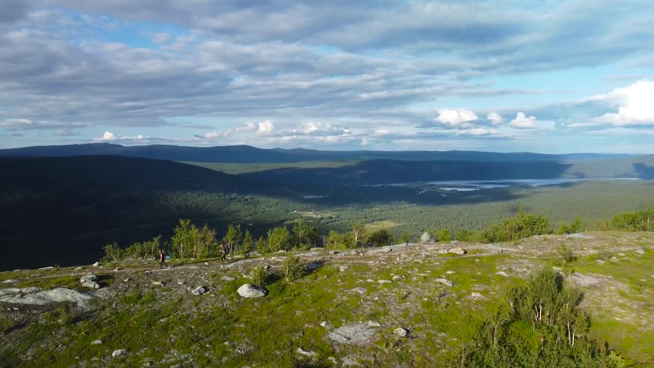 Aerial drone footage flying besides two hikers or backpackers walking or hiking on a mountain hill side or a cliff edge with rocky and grassy landscapes during a sunny warm summer or spring day.