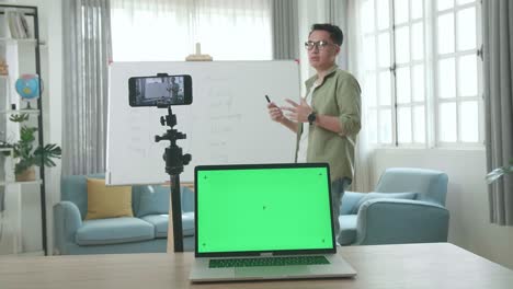 asian male teacher with glasses and green screen laptop shoots video by smartphone camera while teaching english at home