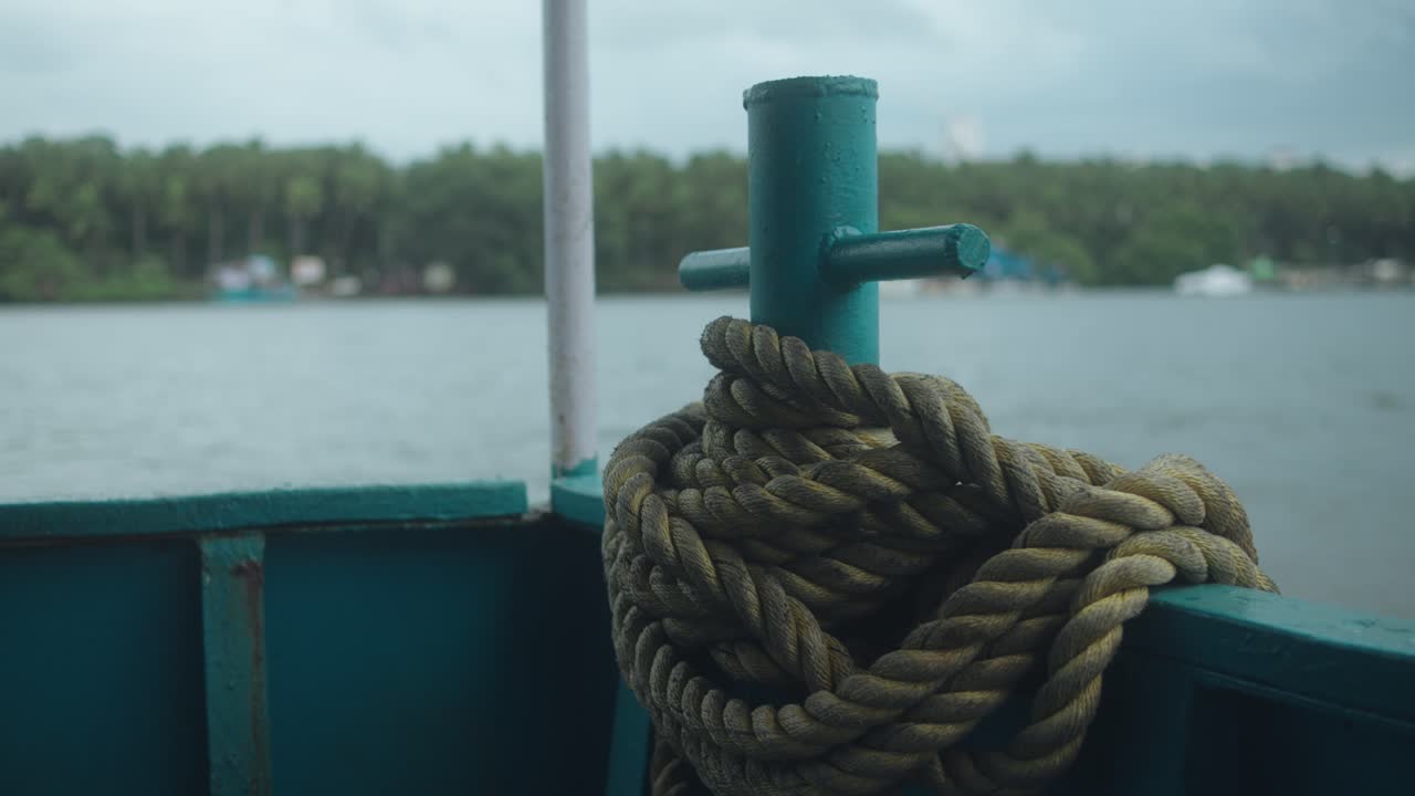 Atmospheric static shot shows a coiled rope on the deck of an old ferry, with a misty river and dense forest lining the distant shore near Old Goa, India