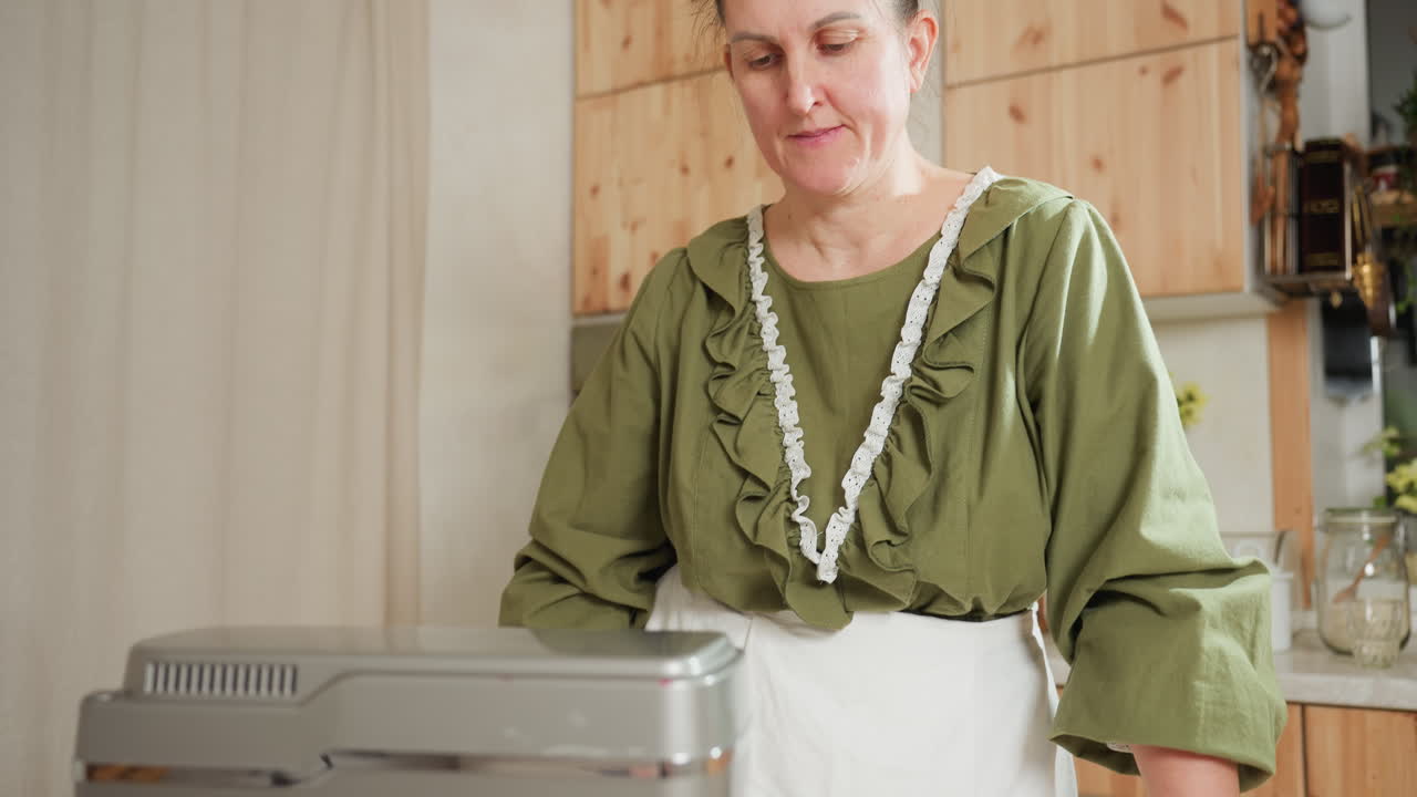 Lady in green ruffled blouse and apron stands calmly with hand on countertop, observing electric mixer in operation during home baking session in modern kitchen with wooden cabinetry
