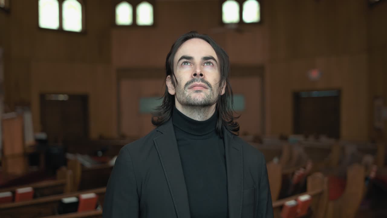 A Christian man, priest, preacher, pastor smiles in a black turtleneck and blazer stands in a church sanctuary, framed by wooden pews and soft light, creating a cinematic and contemplative mood