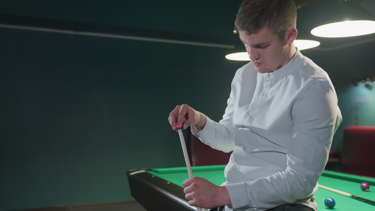 primer plano de un joven con camisa blanca, sentado, sosteniendo un palo de taco y aplicando cuidadosamente tiza roja con expresión enfocada. configuración de sala de billar débilmente iluminada con mesa de billar verde y bolas en el fondo