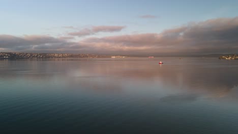 A-single-red-and-white-tug-boat-crosses-calm-waters-as-colorful-clouds-stream-across-the-background,-aerial-timelapse