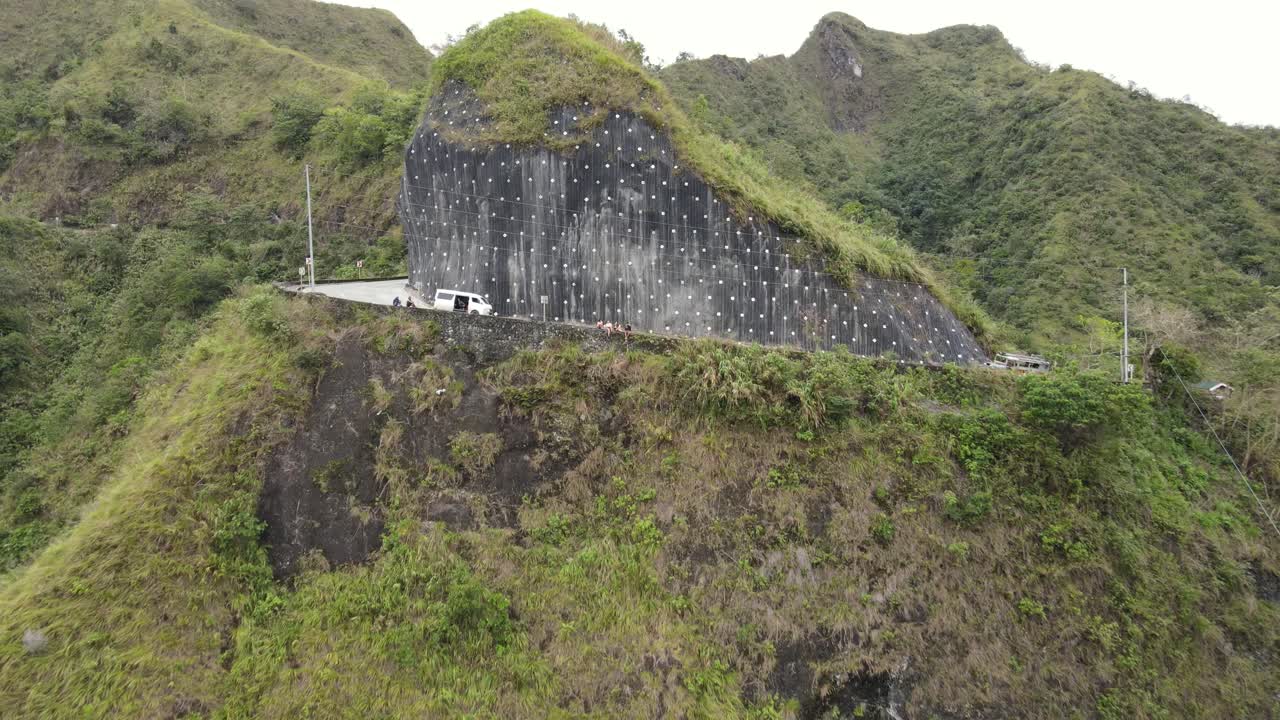 A relaxed group of friends sits along a roadside ledge during a van trip across the majestic Ifugao mountains. The drone pans and turns revealing a winding river in between a deep green valley