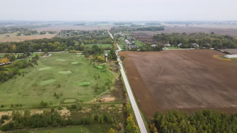 aerial view of countryside nature landscape at cloudy day in autumn season, road through green golf course field and vast agricultural lands, stationary vehicles at roadside, skyline in horizon