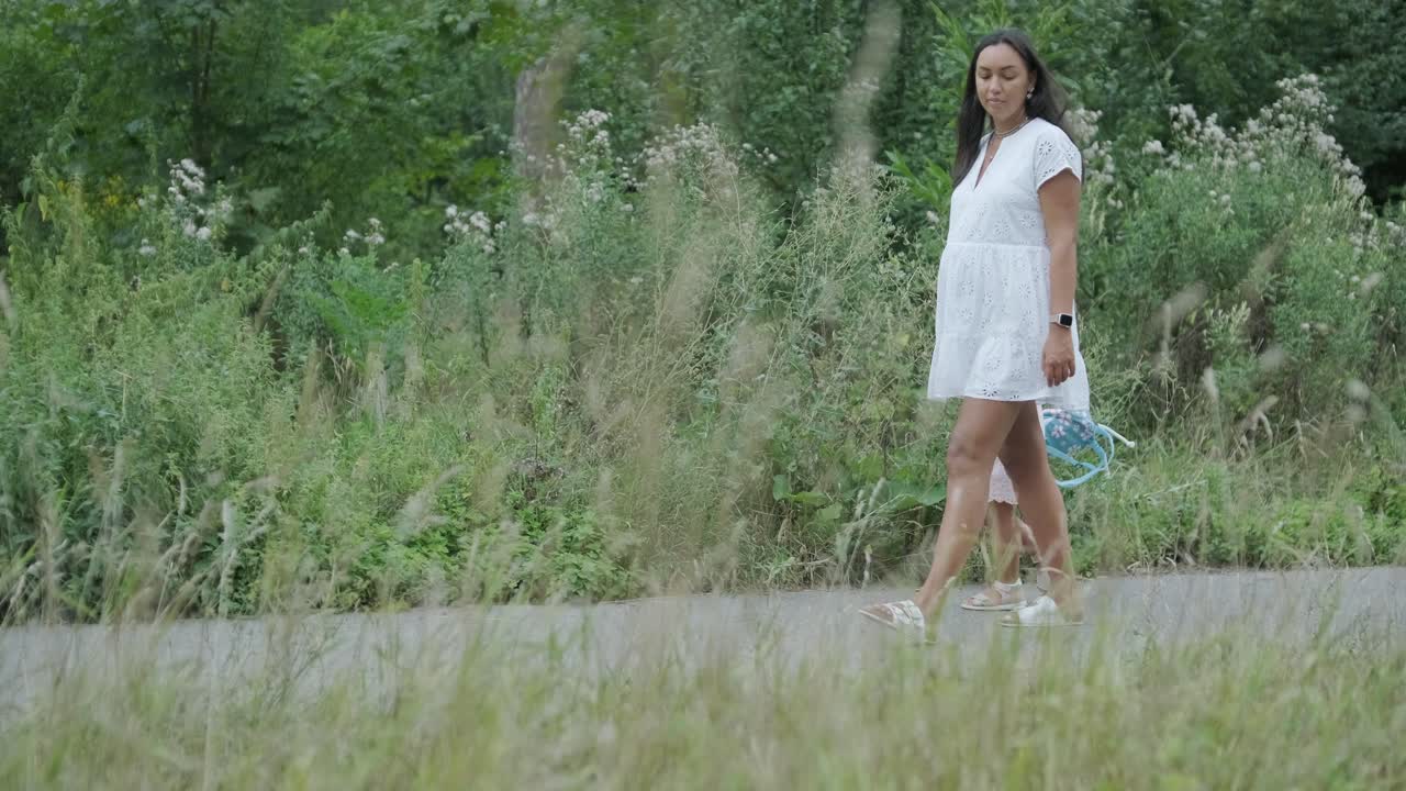 옷을 입은 임신부와 어린 딸이 잔디 인 공원을 가로질러 손을 잡고 걸어가는 장면은 나무로 둘러싸인 자연 속에서 가족의 유대감의 평온한 순간을 포착합니다.