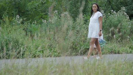 a pregnant woman and her young daughter, both dressed in white, walk hand-in-hand through a grassy park. the scene captures a serene moment of family bonding in nature, surrounded by trees.