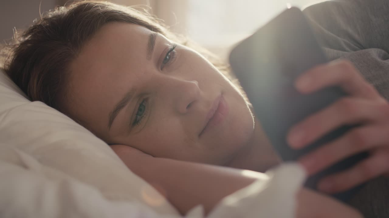 Kaukasisk kvinde ligger i sengen om morgenen og bruger mobiltelefon
