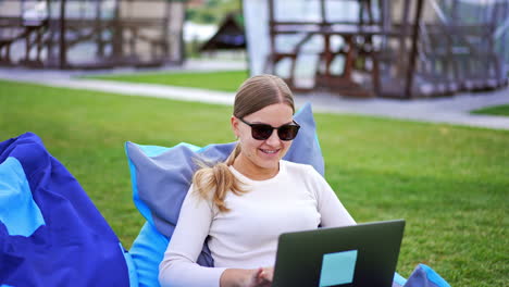 happy relaxed young lady doing her job outdoors. blonde lady in sunglasses sitting in bean bag chair working on laptop. close up.