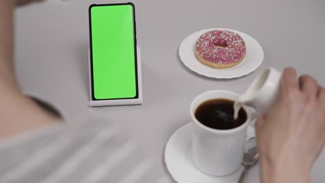 woman sitting at table coffee donut using smartphone with chroma key green screen, scrolling through social network media online shop internet. smartphone in horizontal mode with green screen mock-up.