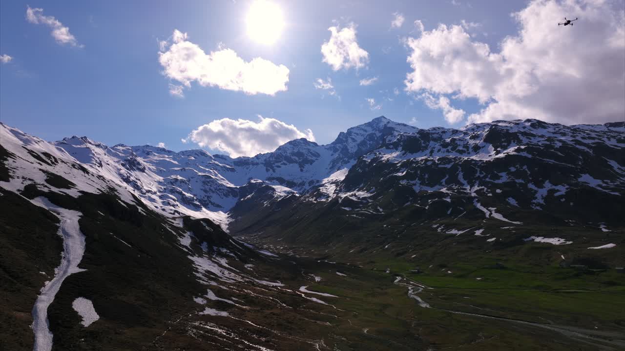 Betagende luftfoto/droneoptagelse af en solbeskinnet alpedal i Montespluga. Viser majestætiske sneklædte bjergtoppe, grønne skråninger og en dramatisk himmel. Italienske Alper, Lombardiet, Italien