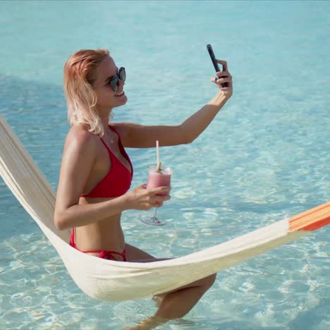 Female-tourist-drinking-cocktail-and-taking-selfie-on-hammock