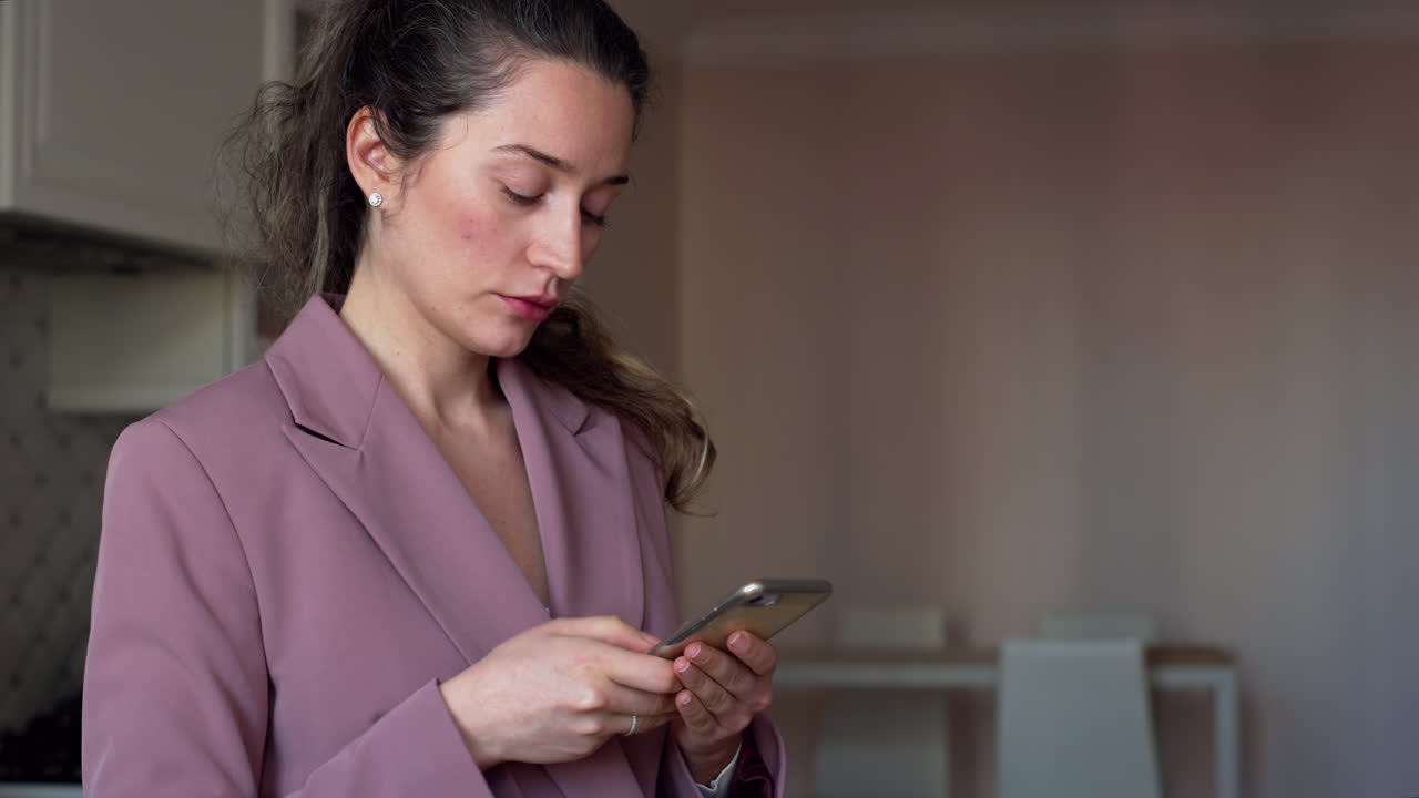 Donna con un blazer rosa in piedi in un ambiente interno che usa il suo smartphone, concentrata sulla lettura o sulla digitazione