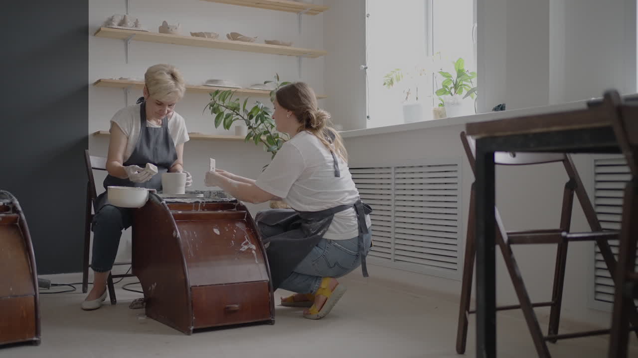 Woman master transfers knowledge to an elderly woman working on a potter's wheel and making a mug of ceramics in her workshop in slow motion