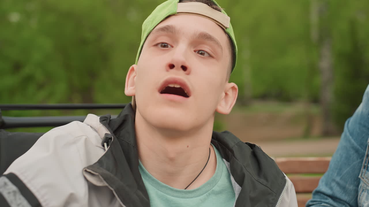 Young Man Playing Happily In Park, Carefree Boy In Wheelchair Having Fun Outdoors With Caregiver Nearby, Energetic Young Man In Wheelchair Interacts Cheerfully With Camera In Sunny Park Setting