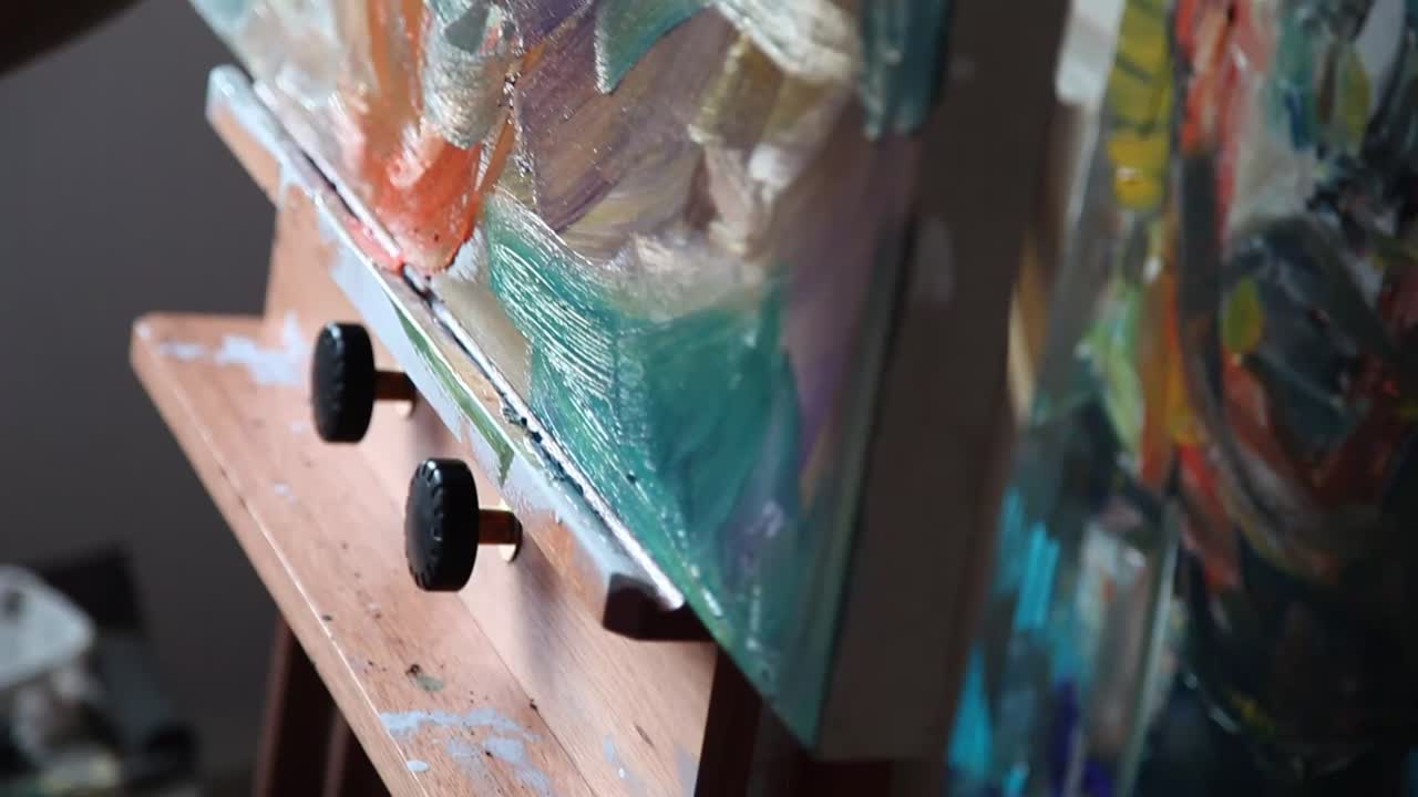 The image shows a tattooed arm holding a paintbrush, creating textured strokes on a canvas. The painting features soft, blended colors like white, blue, and gray, with an abstract and expressive style