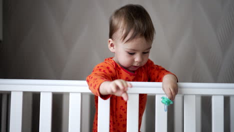 caucasian toddler in orange shirt tries to reach camera with his hand. baby boy standing in the cot drop a blue pacifier and looks how it falls.
