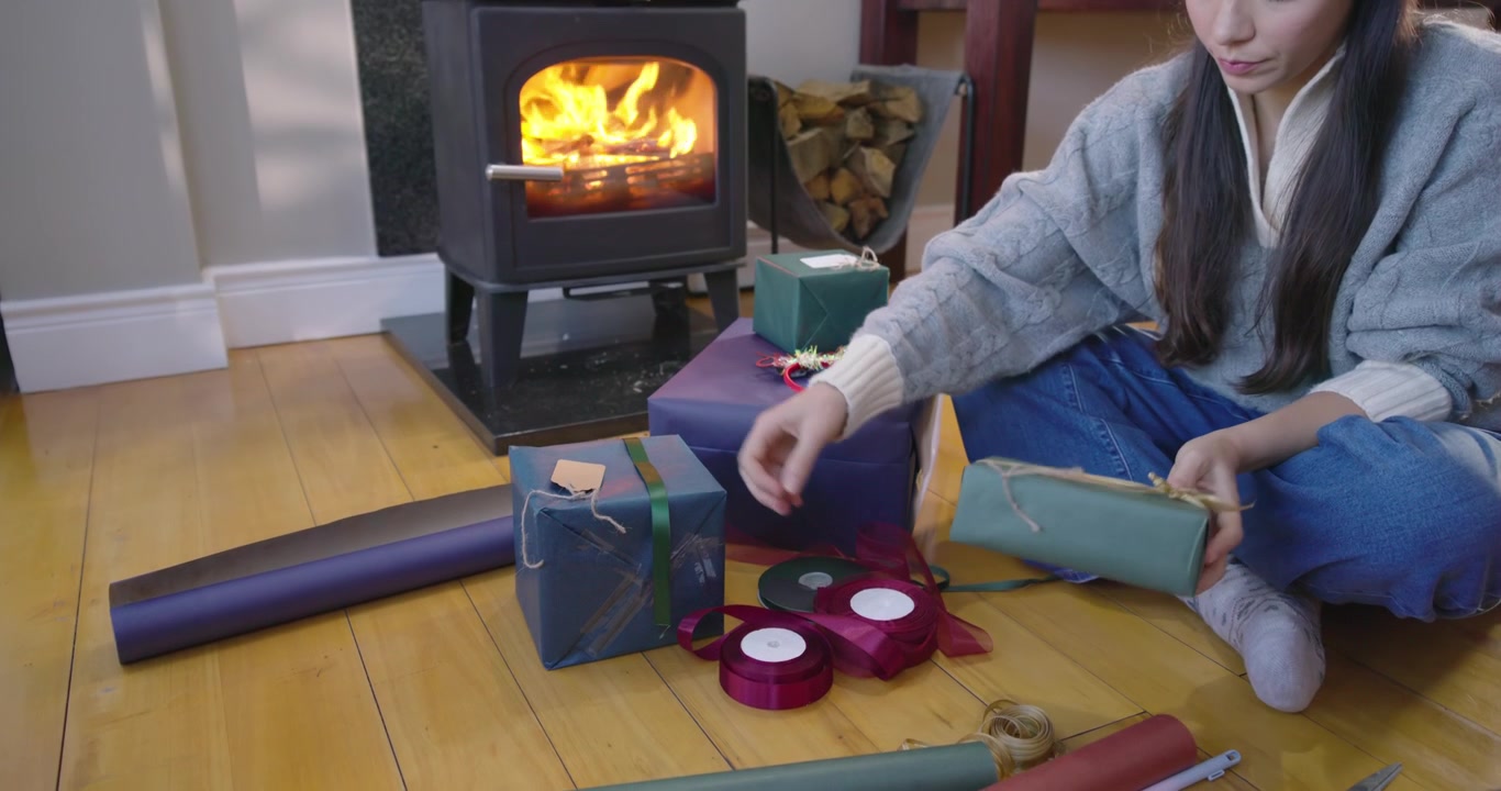 Asian woman sitting by wood stove, preparing gifts, wrapping with ribbons and twine, copy space