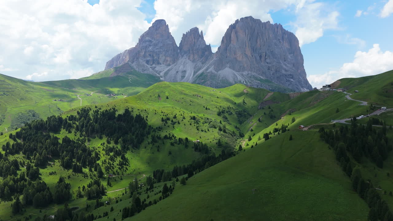 Drone ascends over winding alpine road revealing summer Dolomites with lush green slopes, forests and majestic peaks under moving clouds