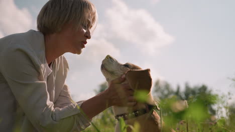 woman holding dog's head lovingly brings face close as dog playfully licks her mouth and his nose in grassy field under bright sky, affectionate interaction showcasing strong bond