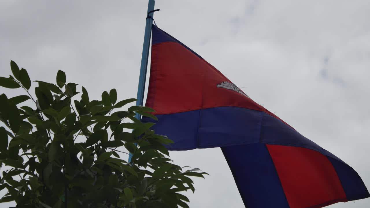 A mid shot of the Cambodian flag waving gently in a natural landscape surrounded by trees and greenery.