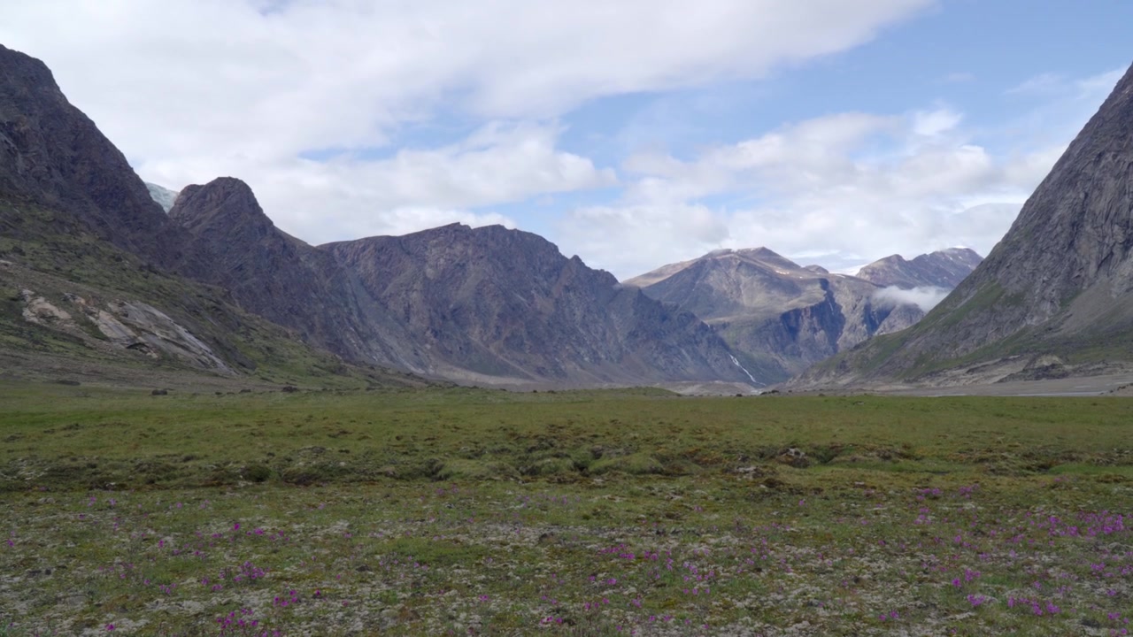 Bredt, symmetrisk bakkenært bilde som ser nedover en vidstrakt tundradal mot taggete fjelltopper under en delvis skyet himmel.