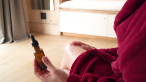woman in cozy robe holding essential oil bottle, relaxing at home