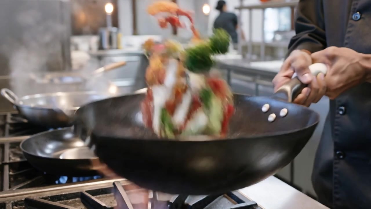 Chef in giacca scura che, afferrando un wok, salta le verdure, cucinando in una cucina commerciale.