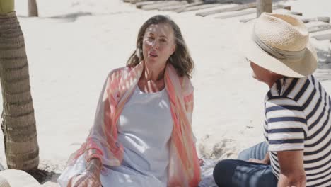 Happy-senior-caucasian-couple-sitting-on-beach-talking-in-the-sun,-in-slow-motion