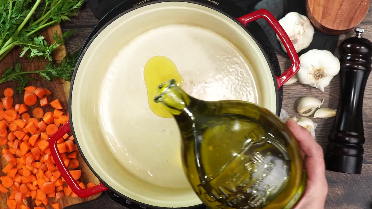Cooking dinner and adding extra virgin olive oil to a red stock pot surrounded by fresh vegetables and herbs. Close up of pouring olive oil into a pot. Cooking a meal and starting with oil to heat up