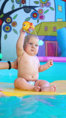 beautiful caucasian baby sits on the mat floating in the pool. kid plays with a toy as coach holds him carefully. vertical video.