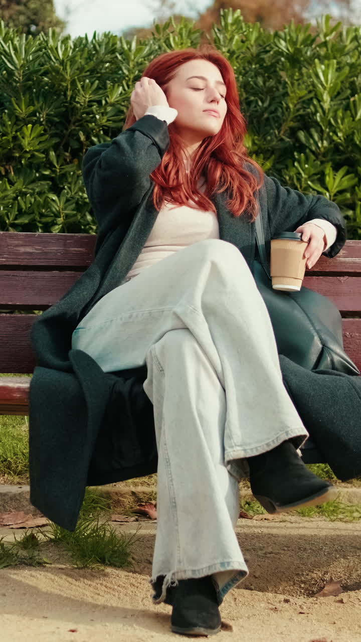 Giovane donna dai capelli rossi si rilassa su una panchina del parco con un caffè