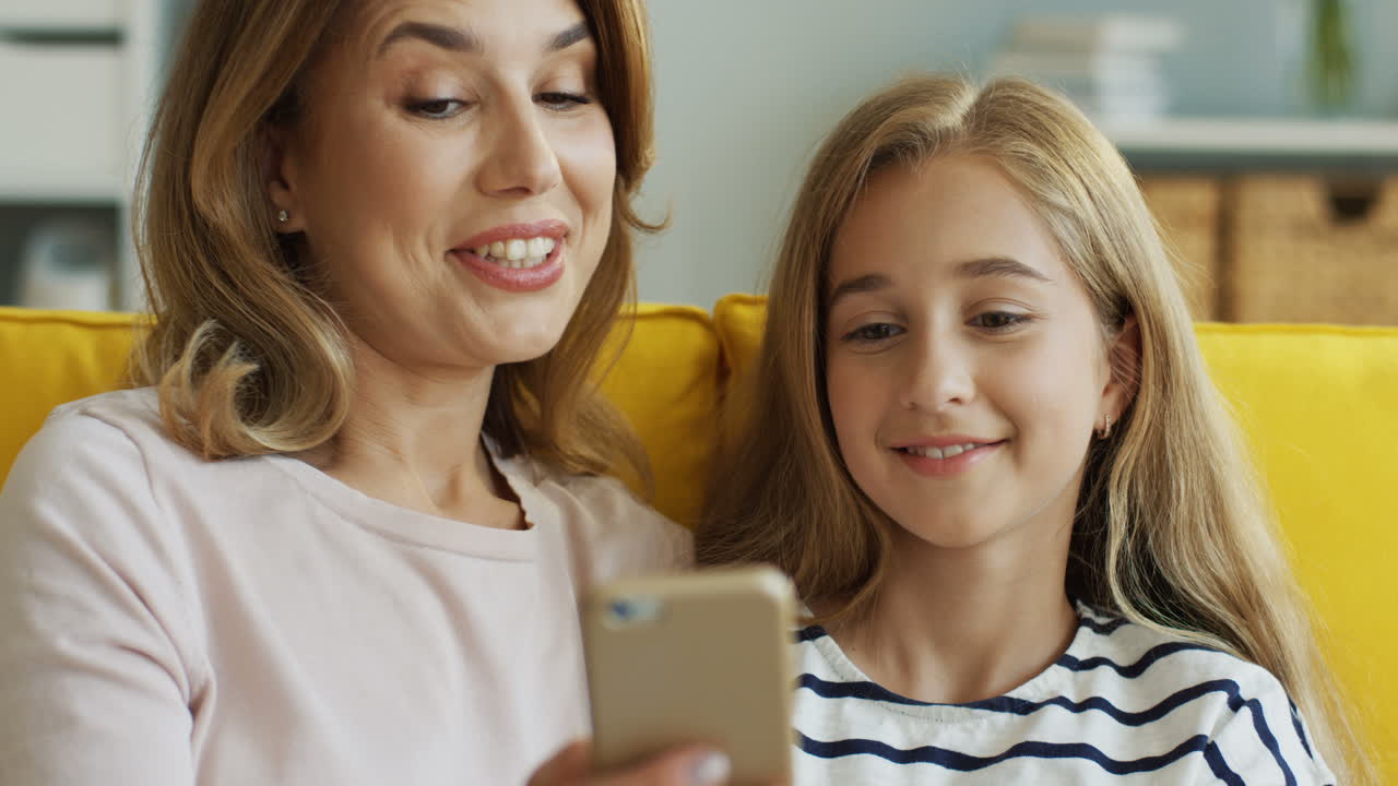 cerca de la madre y la hija caucásicas sonrientes descansando en el sofá en casa y viendo algo en el teléfono inteligente y hablando. interior.
