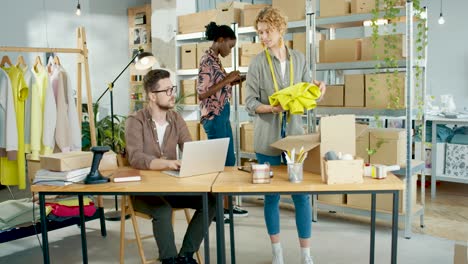 multiethnic designers team browsing online on laptop, packing clothes, typing on tablet and checking boxes in fashion clothing shop