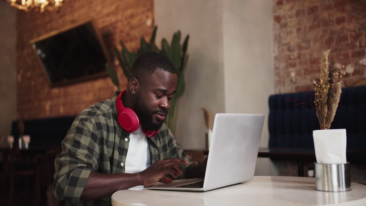 A black character with a beard in red headphones emotionally works on a laptop in a cafe. Video filmed in high quality