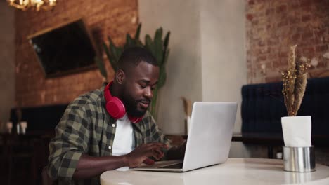 A-black-character-with-a-beard-in-red-headphones-emotionally-works-on-a-laptop-in-a-cafe.-Video-filmed-in-high-quality