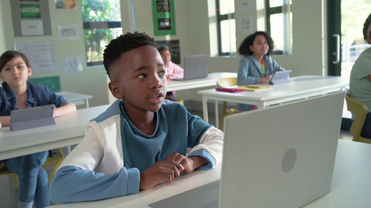 Ragazzo afroamericano alla scrivania che alza la mano, rispondendo all'insegnante, usando un laptop argentato
