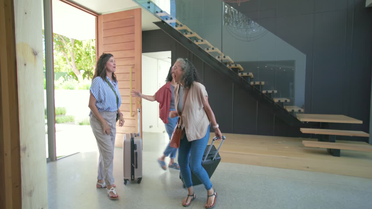 Diverse women friends entering foyer as guests, hand pulling door with bags, gazing at chandelier
