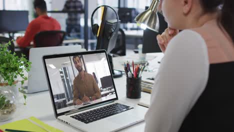 mujer de negocios caucásica sentada en un escritorio usando una computadora portátil haciendo una videollamada con un colega masculino