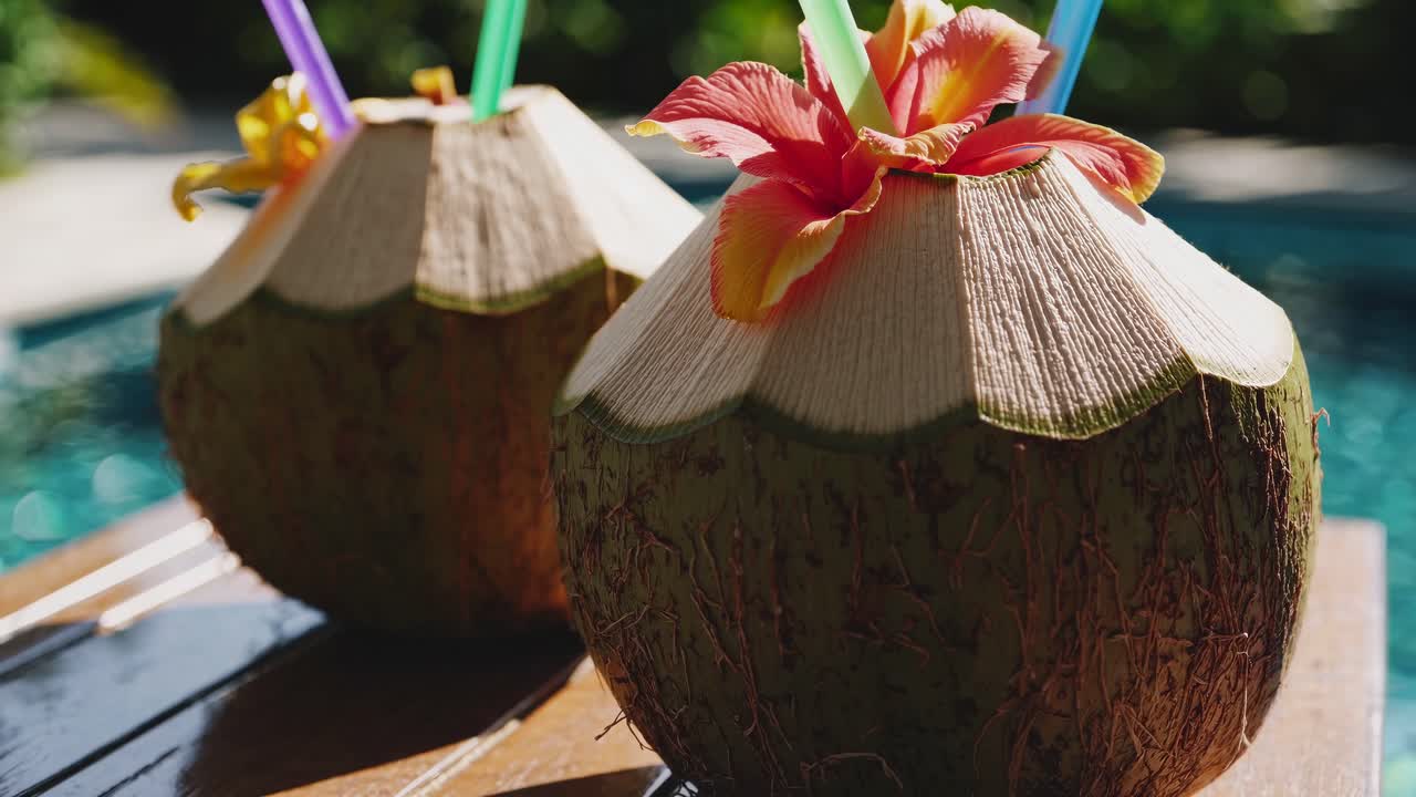Close-up, eye-level view of tropical coconuts with colorful straws and flowers by a pool